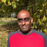 Headshot of London SLC member, Haren Thillainathan. He standing outside under a tree in the sunshine. He is wearing a red and navy t-shirt.