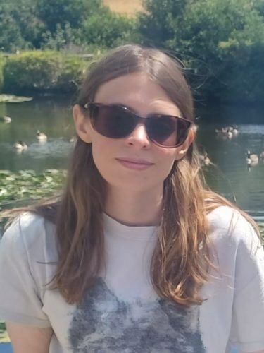 Image of Lauren Eade, South East SLC's coordinator. Lauren is sitting in a garden in the sun. She has long, brown, hair and is wearing sunglasses.