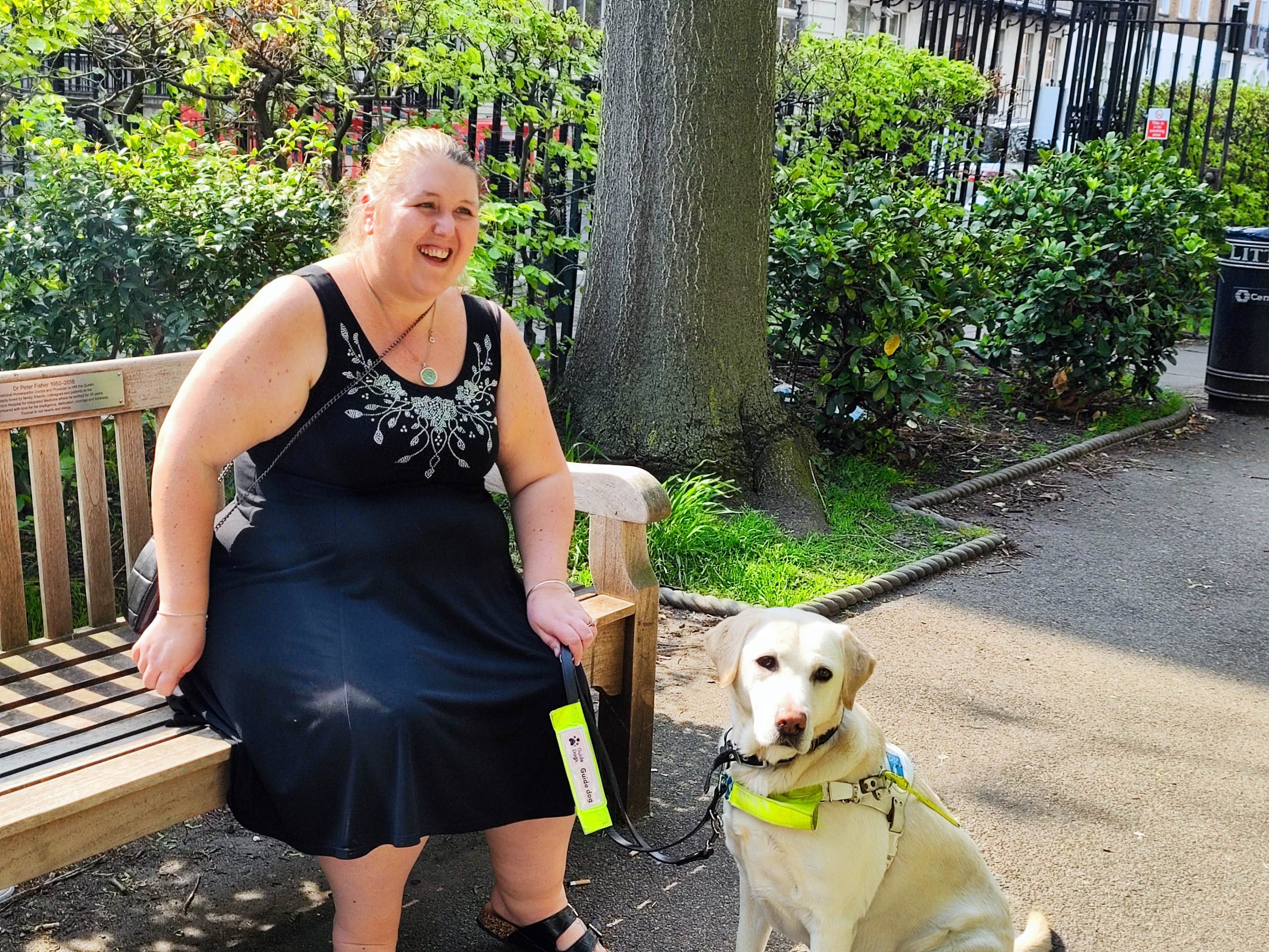 Leanne, London SLC member is sitting on a park bench with her guide dog. She is looking forwards, laughing.