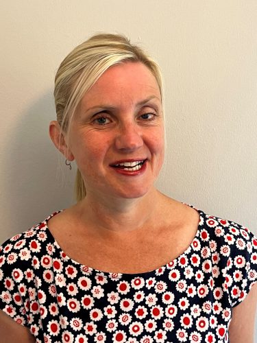 headshot of Kelly Barton, Engagement Manager for the north west. Kelly is standing against a white wall and smiling at the camera. Her hair is pulled back into a pony tail. She is wearing a black top with red and white flowers on it.