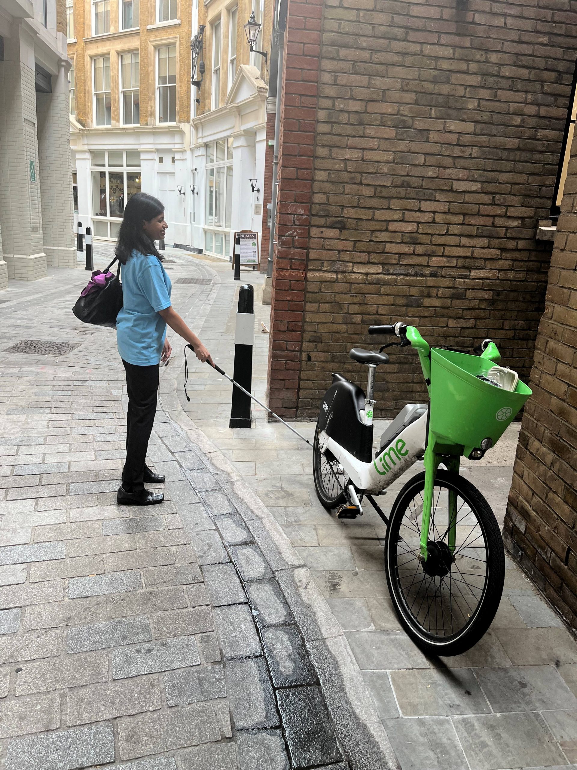 Vidya Nathan, London SLC member, is crossing a road. Using her long cane, she encounters a badly parked Lime Bike, obstructing the entrance to an alley way that she wants to walk down.