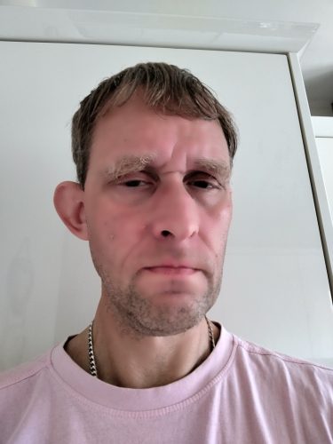 Headshot of ashley Pearce, SW London SLC member. He is standing against a white wall, wearing a pale pink t-shirt. He is looking down at the camera.