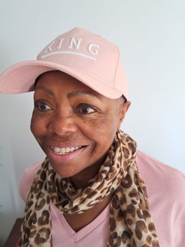 Lola, South East London SLC member is wearing a baby pink baseball hat and sweater, and a leopard print scarf. She is looking away from the camera, with a big smile on her face.