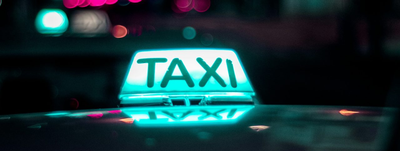 Close up of a taxi light, lit up in blue and white. The background is dark - night time - with some twinkling lights shown in the background.