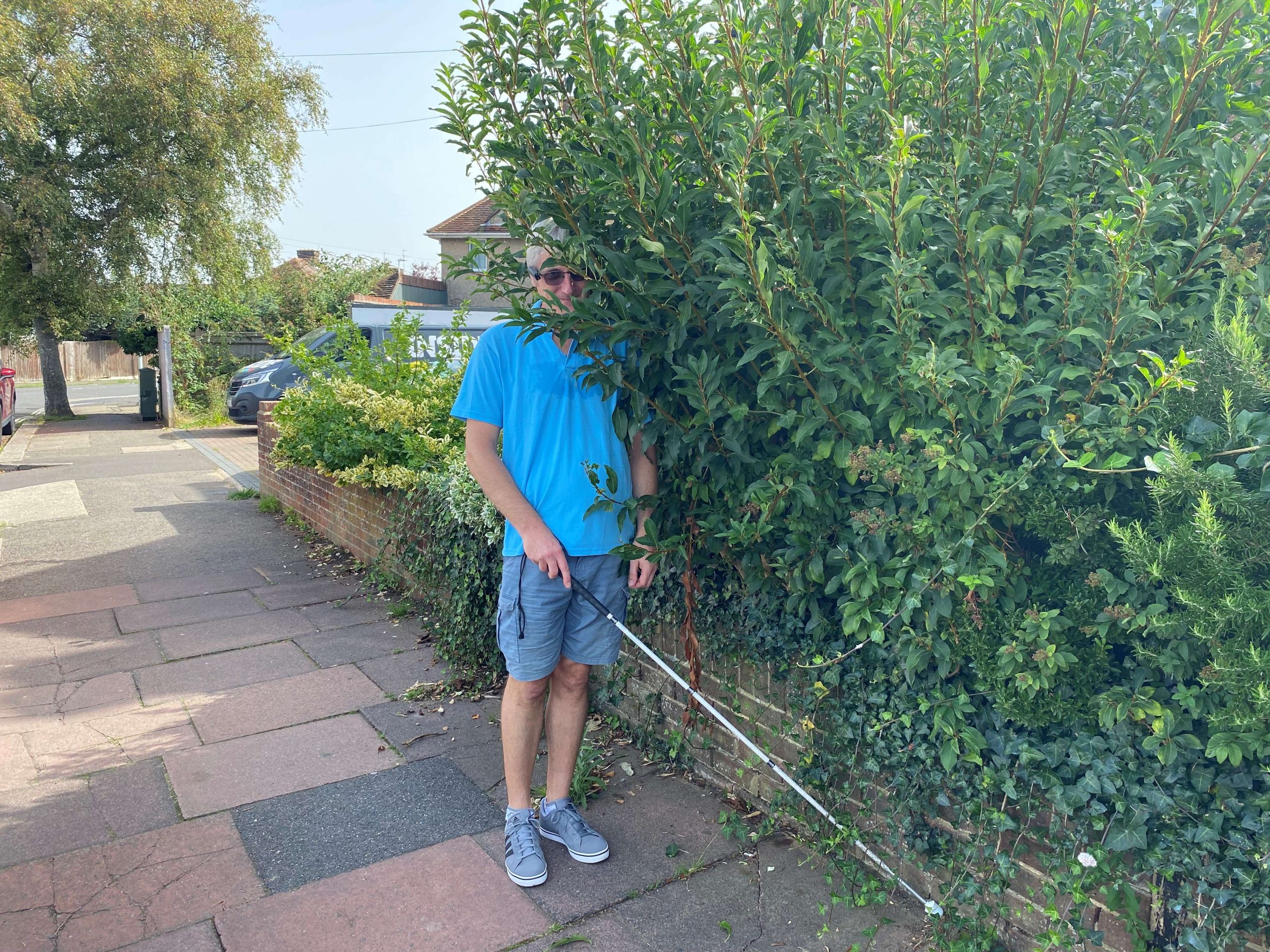 West Sussex SLC member, Clinton is shown walking with his long cane, into a large garden hedge which is protruding onto the pavement.