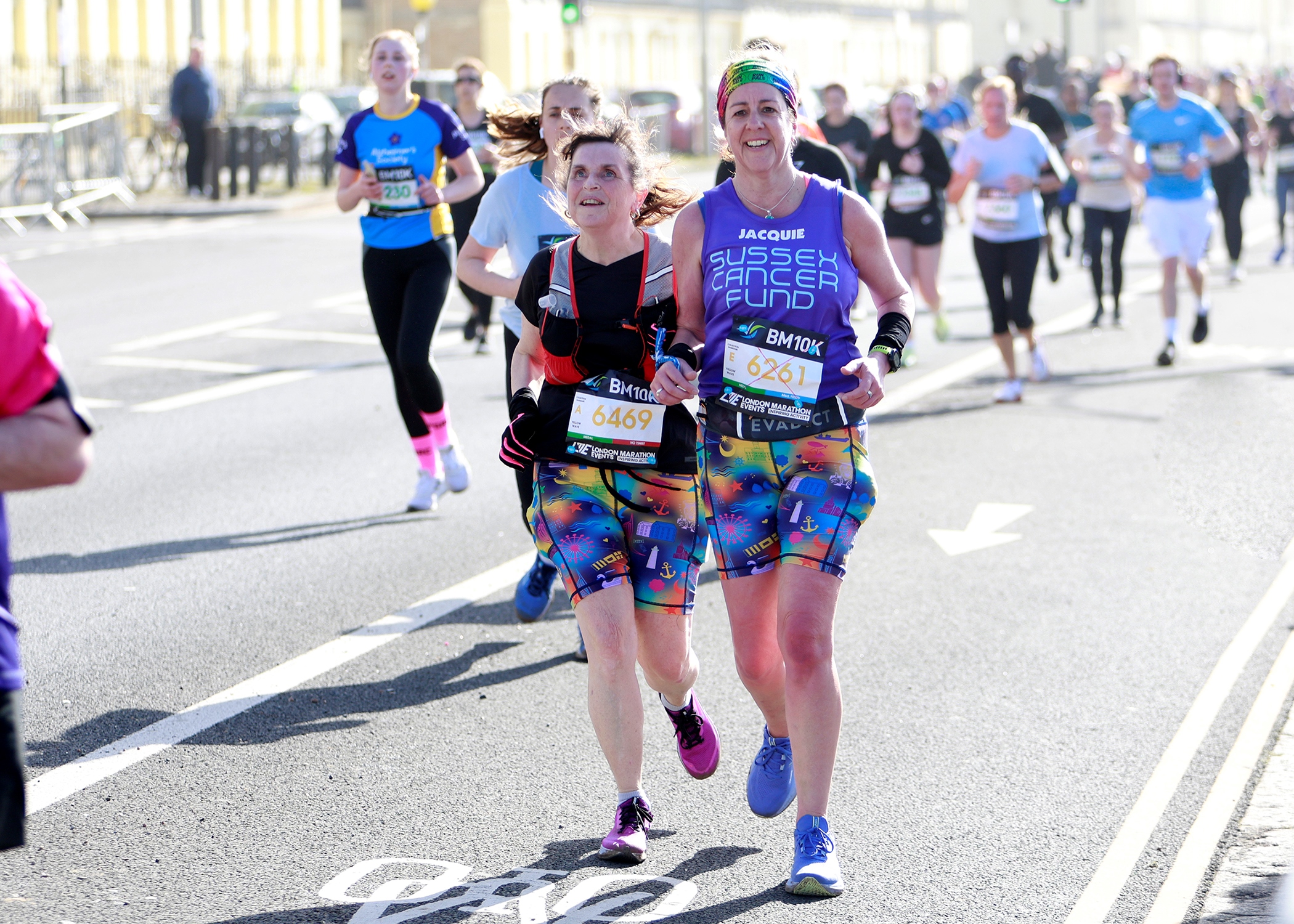 Alison Evans, East Sussex SLC member, pictured running with a guide runner. She is wearing a race bib, and is pictured running down a main road with other runners shown in the background.