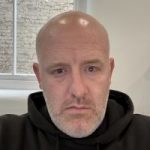 Headshot of Josh Brown, Nottinghamshire SLC member. Josh has a shaved head and a short, grey beard. He is wearing a black hoodie and looking at the camera.