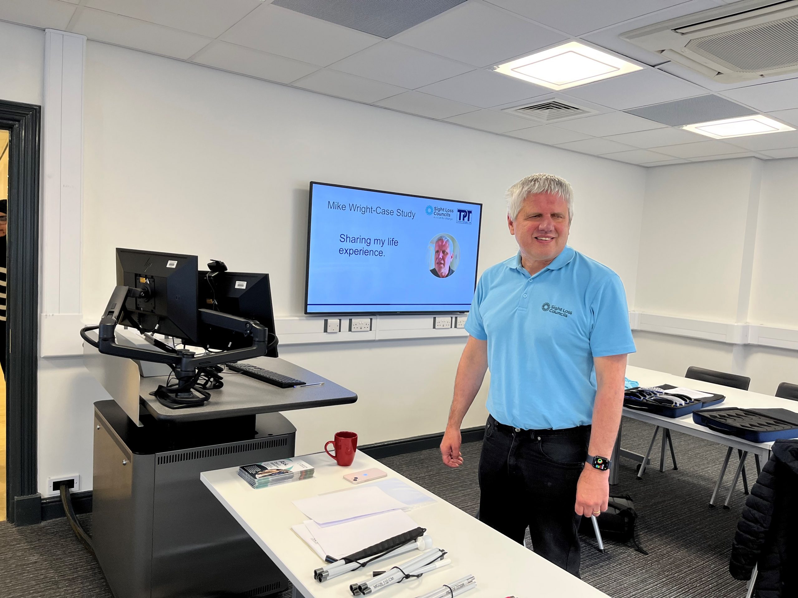 Mike Wright, Merseyside SLC volunteer, standing behind a table during his presentation in a classroom. His head is turned to the side and he is smiling. He is wearing a pale blue Sight Loss Council t-shirt, and in the background is an overhead projector with his name and photo on it.