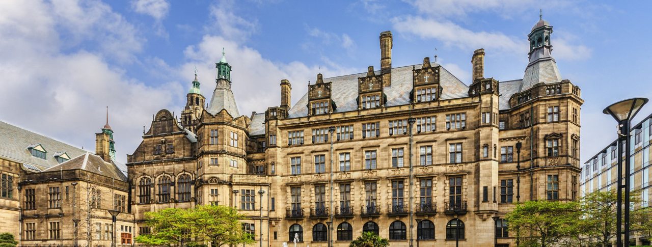 Landscape image of Sheffield Town Hall, a Grade 1 listed building built with Stoke stone and ornate carvings. In front of the town hall is a circular fountain, grass and trees.