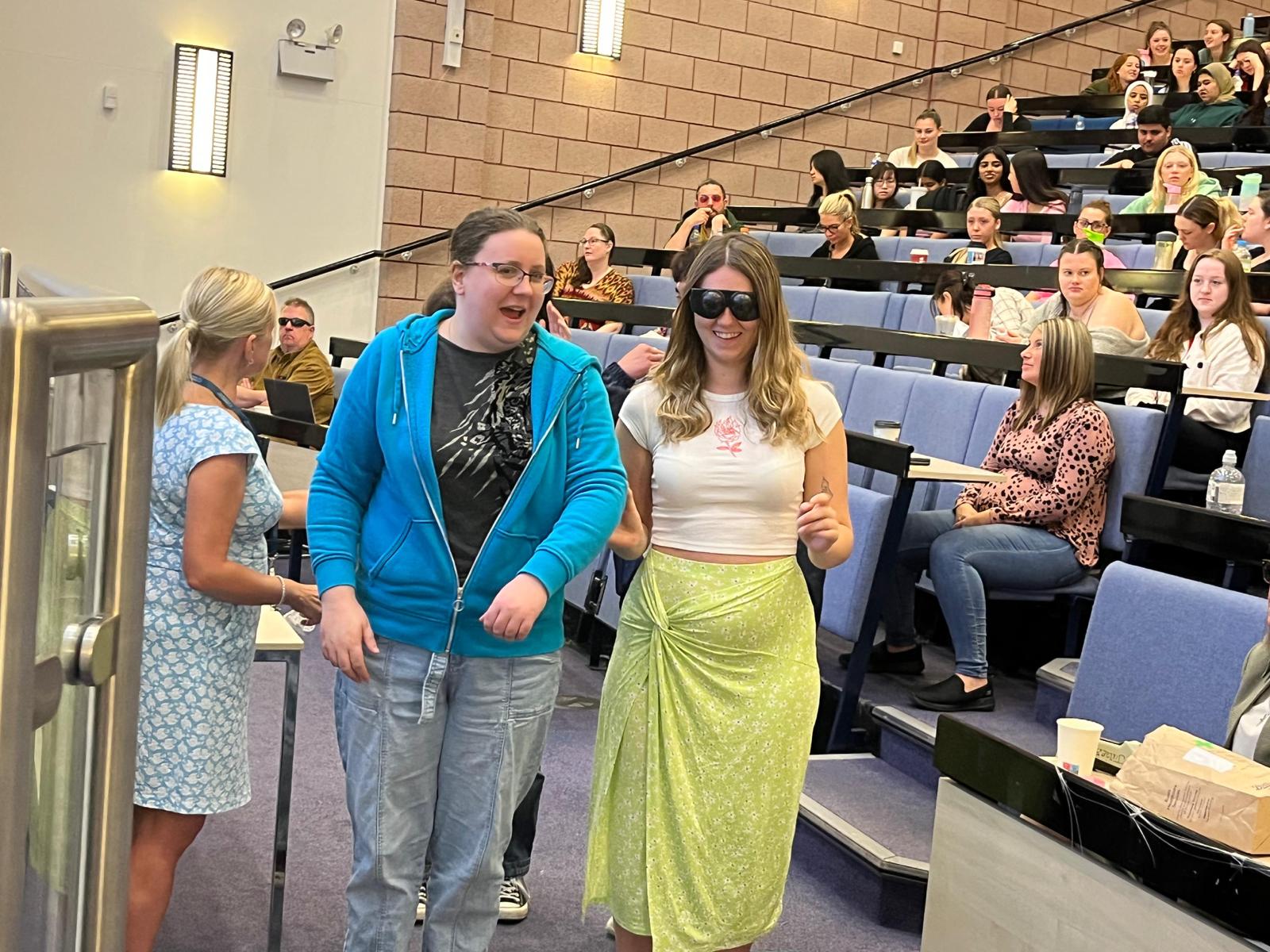 Two female university students during the sighted guide session in the auditorium. Kelly Barton, Engagement Manager for northwest England, is stood to the left hand side of them.