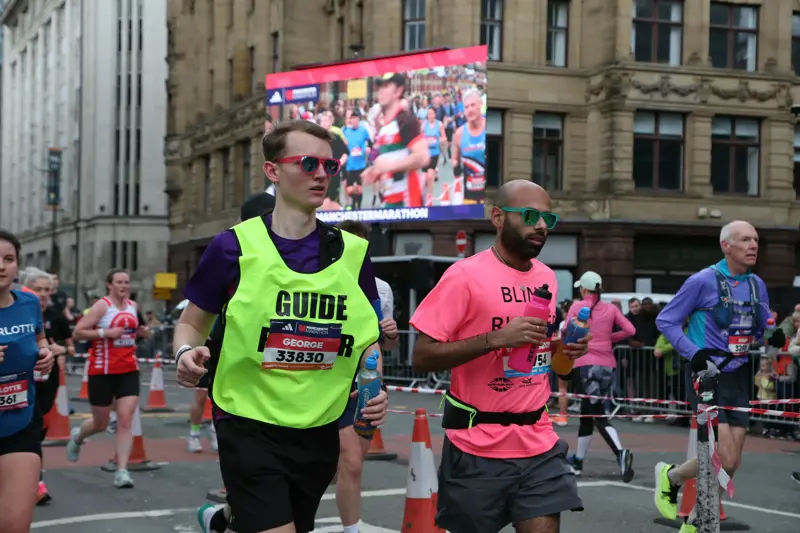 ahya running the Manchester Marathon next to his guide runner, George. Yahya is wearing a neon pink t-shirt which says 'Blind runner' on it, George is wearing a neon yellow bib which says 'Guide runner' on it.