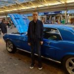 Jordan Brennan, South East London SLC member. Jordan is standing in front of a vintage blue car which has it's bonnet up.