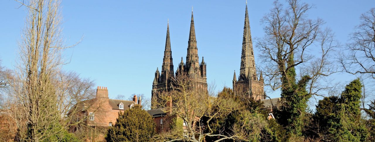 Landscape image of Lichfield Cathedral in Staffordshire. The photo is taken from the other side of the river. Two people are sat on a wooden park bench in front of the cathedral, and large trees frame the cathedral.