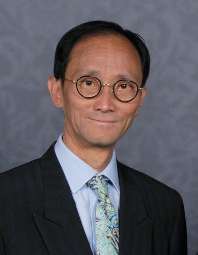 Paul Hsu, North London SLC member. Paul is wearing a dark suit and stood against a grey background. He is wearing glasses, and smiling at the camera.