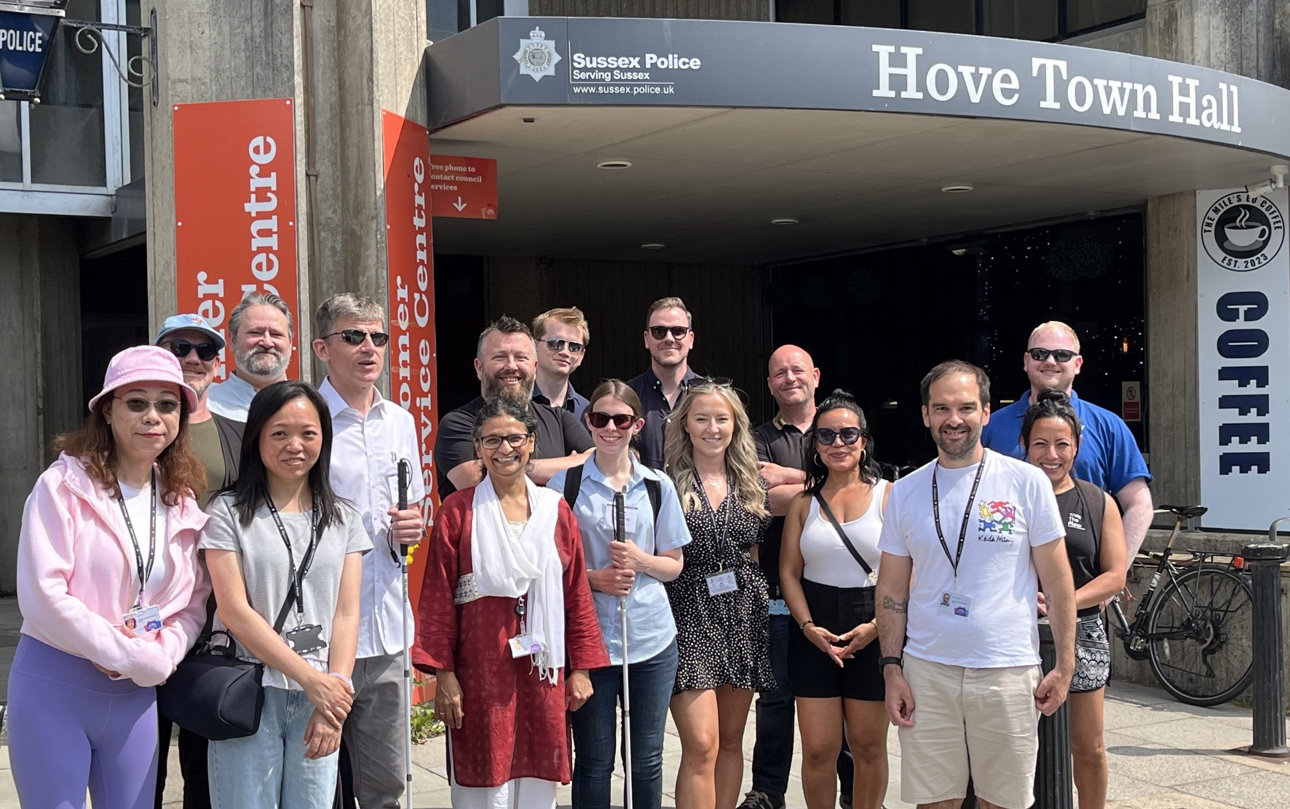 A group shot of 16 Sight Loss Council (SLC) members and council officers stood in a line smiling at the camera infront of Hove Town Hall, with SLC Engagement Manager Dave Smith and SLC Co-ordinator Lauren Eade. They are smiling at the camera.