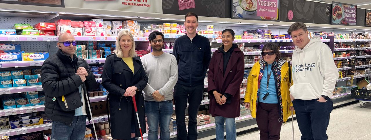 Birmingham and Black Country Sight Loss Council members at a supermarket where they are carrying out market research and giving feedback on packaging for accessibility. They are stood in a line smiling at the camera infront of a dairy isle. Two members are holding a cane. One member is wearing a hoodie with the ‘Sight Loss Councils’ logo.