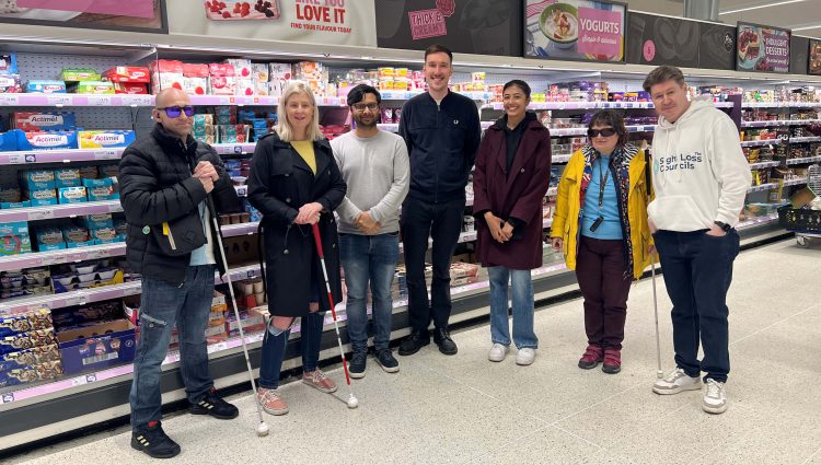 Birmingham and Black Country Sight Loss Council members at a supermarket where they are carrying out market research and giving feedback on packaging for accessibility. They are stood in a line smiling at the camera infront of a dairy isle. Two members are holding a cane. One member is wearing a hoodie with the ‘Sight Loss Councils’ logo.