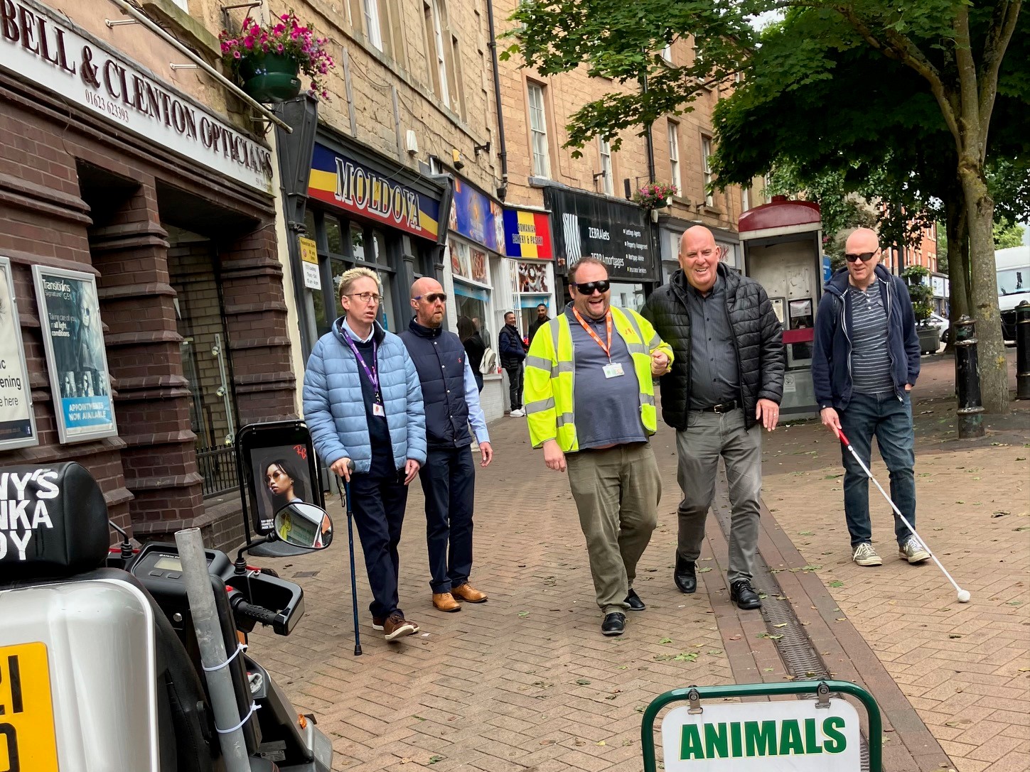 Staff from Mansfield Council being guided down a walkway in Mansfield by Sight Loss Council members whilst wearing simulation spectacles. Street obstacles include an advertising boards and a moped parked on the walkway.