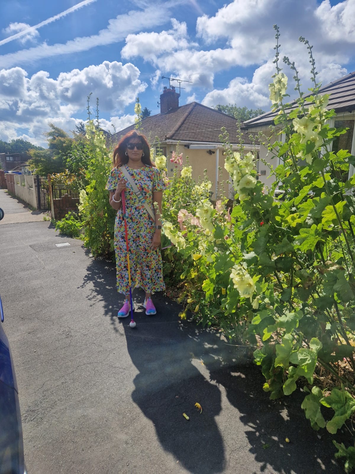 Sight Loss Council volunteer Anela - a visually impaired woman in a long dress and pink shoes, holding a cane, standing on a pavement next to overhanging foliage partially obstructing the path. There is a also the edge of a car on the other side of the pavement further obstructing the path. 