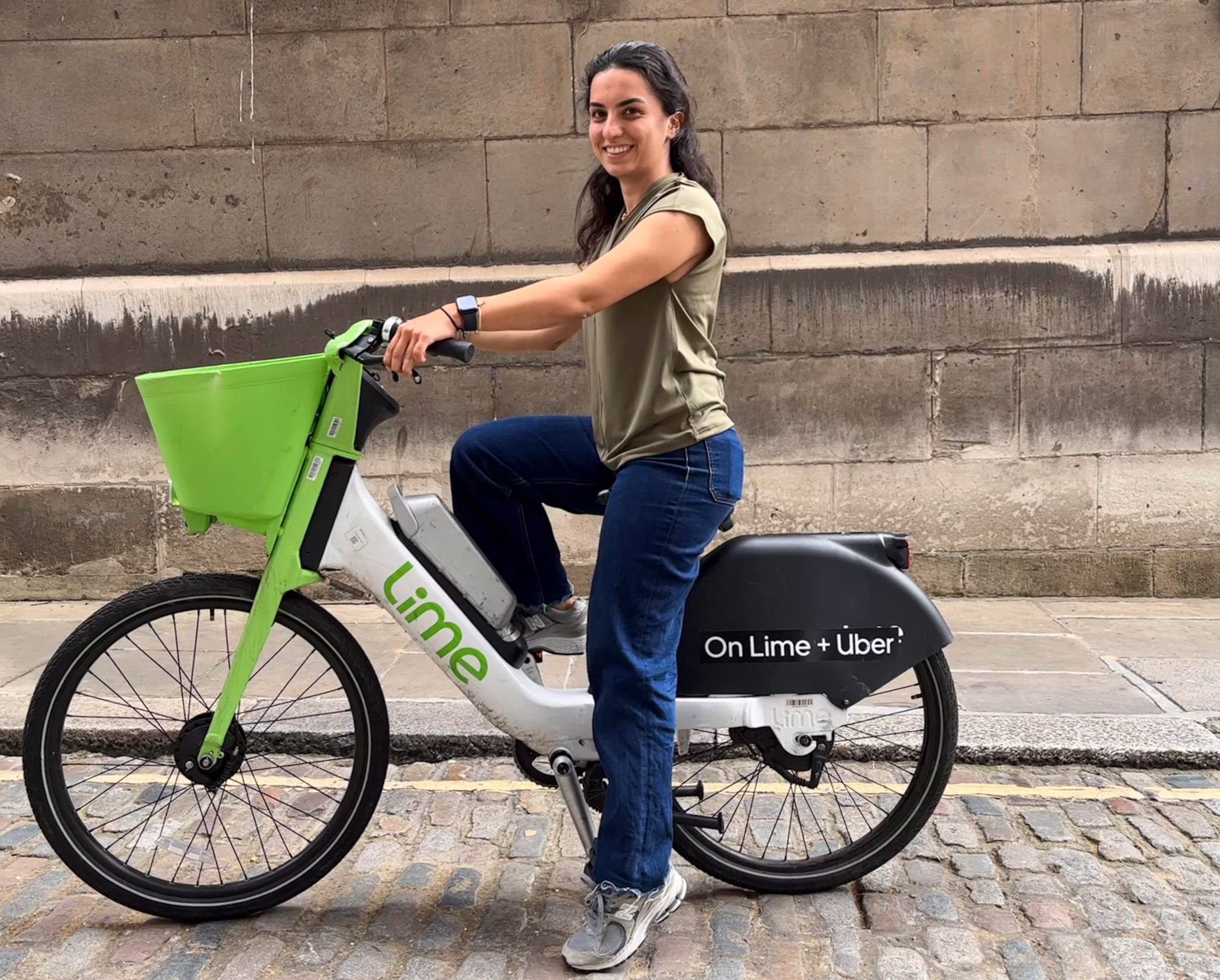 Fiona Duenyas, Public Affairs Manager at Lime, sitting on a stationary Lime bike on a cobbled street. She is smiling at the camera.