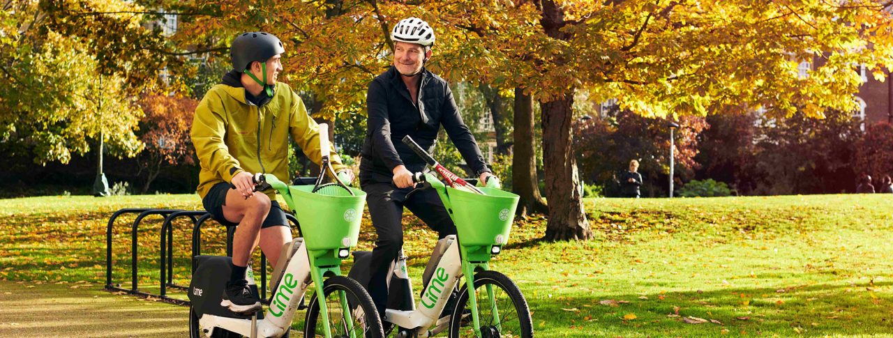 Two people sat on Lime e-bikes on a path. Greenery and trees in background.