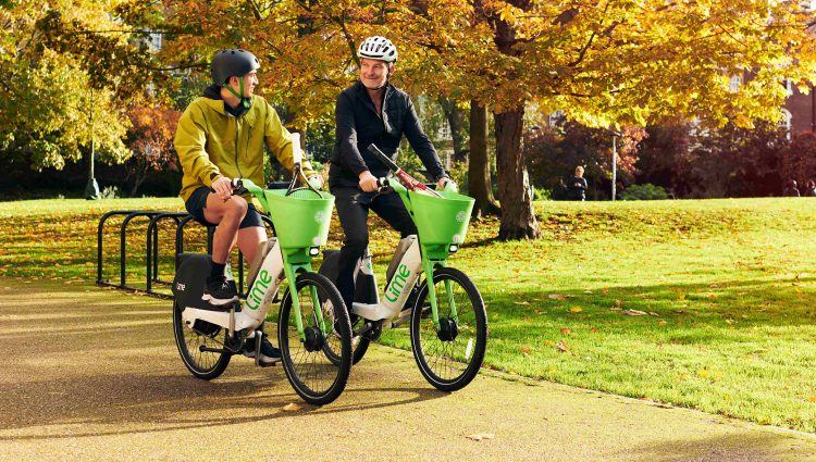 Two people sat on Lime e-bikes on a path. Greenery and trees in background.