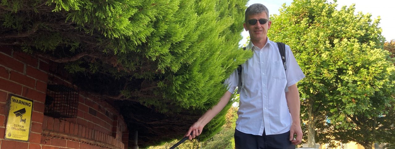 Dave Smith, Engagement Manager for South East England, is pictured next to a large hedge which is overhanging into the pavement. Dave is holding his long cane and looking at the camera.