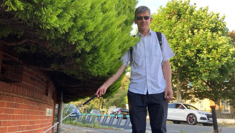 Dave Smith, Engagement Manager for South East England, is pictured next to a large hedge which is overhanging into the pavement. Dave is holding his long cane and looking at the camera.