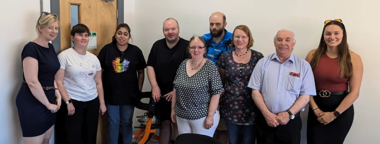 Image shows 7 Staffordshire SLC volunteers standing in a row in front of a white wall and smiling at the camera with Senior Sight Loss Council Engagement Manager Louise Connop (far left) and Sight Loss Council Co-ordiantor Ashley (far right).