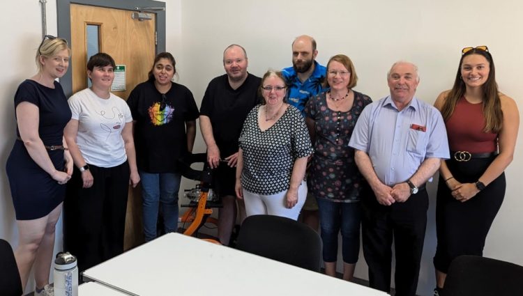 Image shows 7 Staffordshire SLC volunteers standing in a row in front of a white wall and smiling at the camera with Senior Sight Loss Council Engagement Manager Louise Connop (far left) and Sight Loss Council Co-ordiantor Ashley (far right).