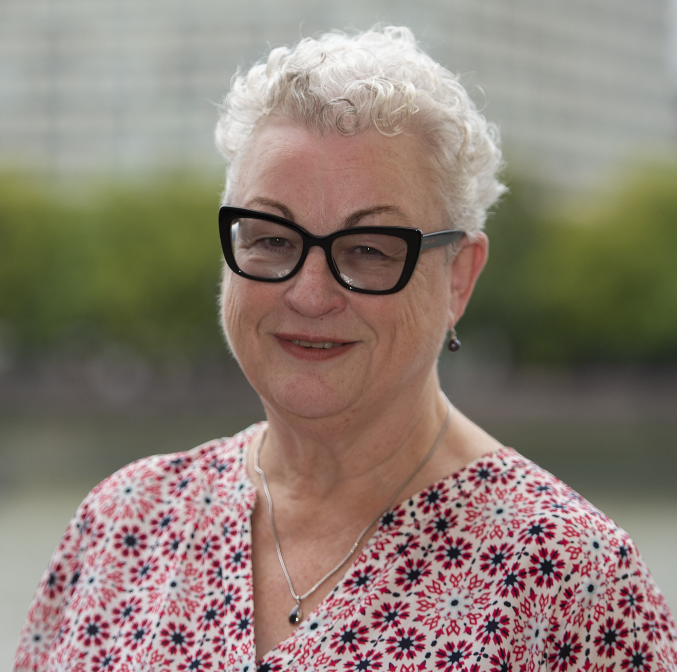 [Headshot of Claire Walters. Claire has white short hair, is wearing a pink floral top and black framed glasses. There is foliage behind her, which has been blurred slightly.]