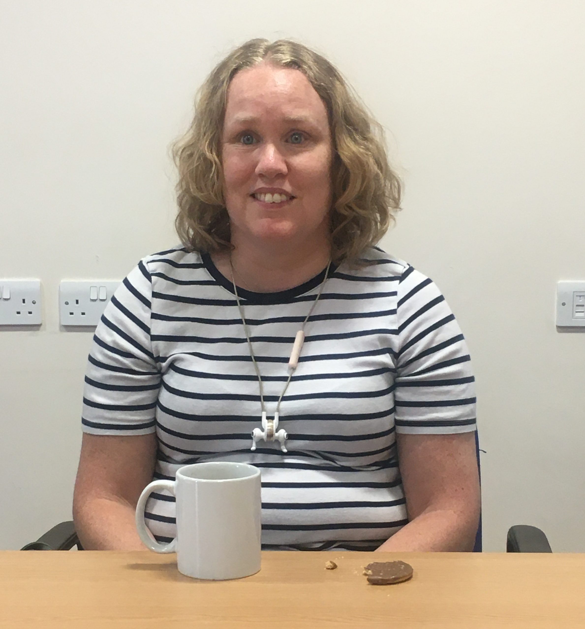 Linn Davies, East Sussex SLC volunteer is sitting at a table with a white mug in front of her. She is wearing a black and white striped t-shirt and smiling at the camera.