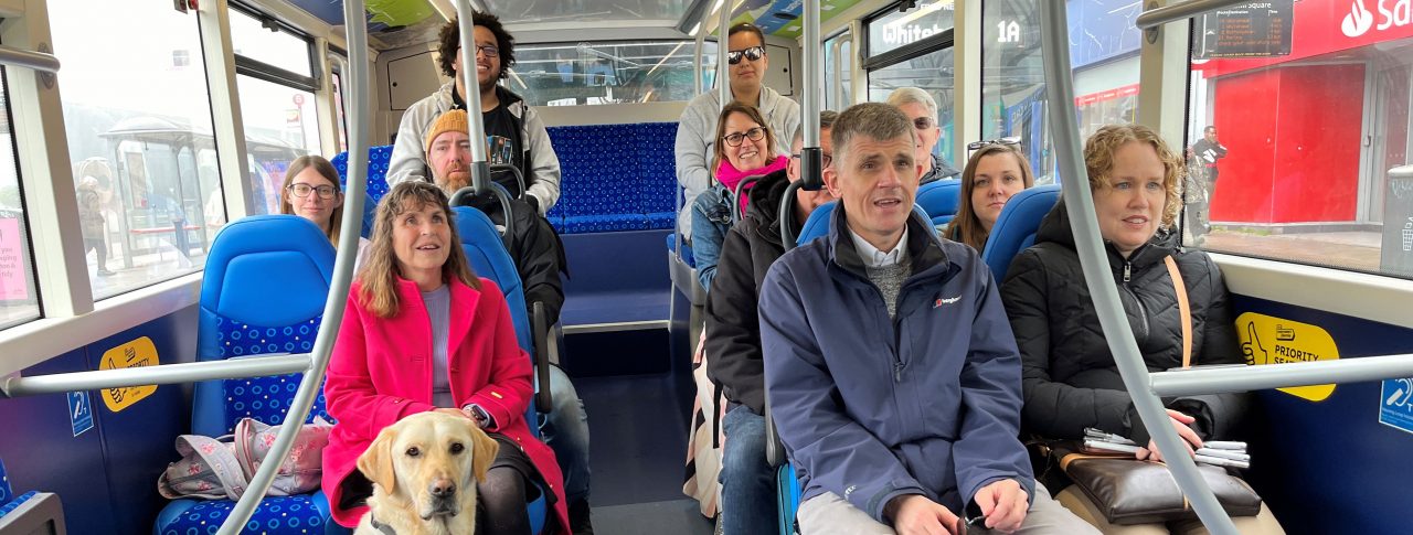 Sight Loss Council and Young Voices volunteers, and Thomas Pocklington staff, are sat on a bus creating some of the videos used in the training materials. Two passengers have white canes with them and there is a guide dog sat at the front.