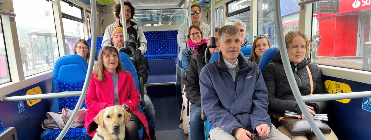 [8 people, including SLC staff members and volunteers sitting on a bus with blue seats and smiling at the camera.]