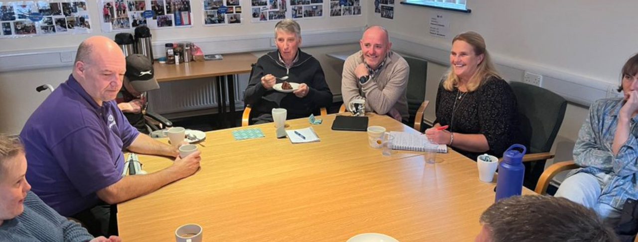 Image shows Hampshire SLC volunteers sitting around a table, with some eating cake. There are photos on the wall behind them and a grey banner with 'congratulations' written in light grey.