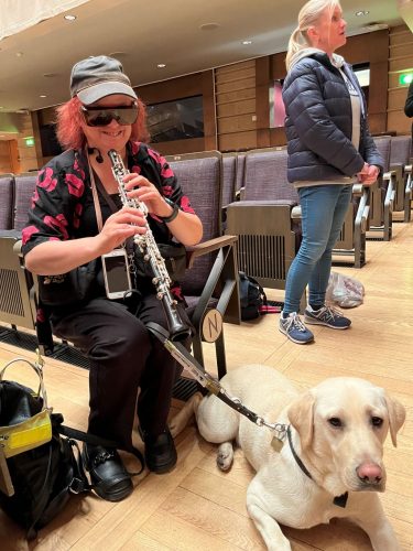 Greater Manchester SLC volunteer holding the clarinet and smiling, facing the camera. Her guide dog sits by her side.