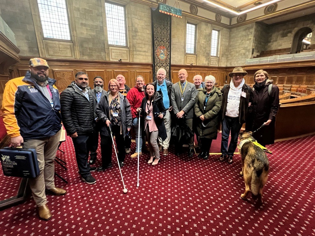 Leeds Councillors stood in a line with West Yorkshire Sight Loss Council members in a large carpeted room in a council building (civis hall). The room has stone walls, with wooden panelling at the bottom, and several tall windows. Two Sight Loss Council members are holding navigation canes and one has a guide dog.