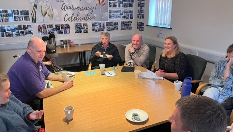 Image shows Hampshire SLC volunteer sitting around a table with some eating cake. There are photos on the wall behind them and a banner with 'congratulations' printed in light grey.