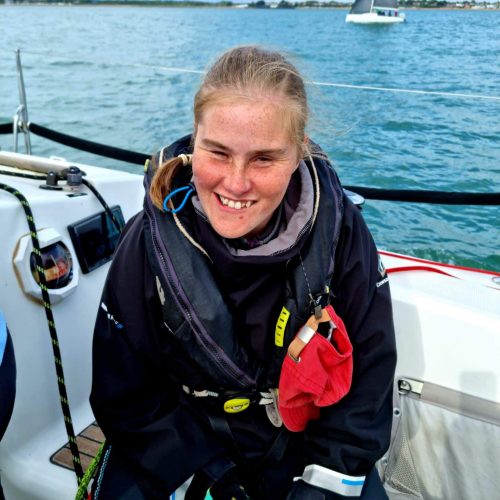 Image shows a headshot of Kate Healey. Kate is sitting on a boat, wearing a black coat and navy life jacket and smiling at the camera. 