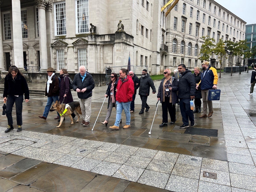 Leeds Councillors on a guided walk on the streets in Leeds with West Yorkshire Sight Loss Council members. Councillors are wearing sim specs. One SLC member has a guide dog, and two are walking with navigation canes.