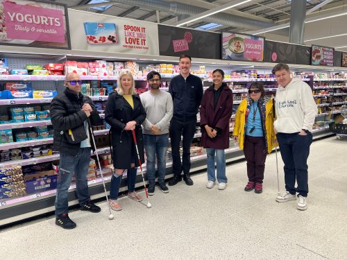 Sight Loss Council members stood in a line with a retailer facing the camera and smiling in a supermarket where they have been assessing the accessibility of product packaging. Three members are holding navigation canes.