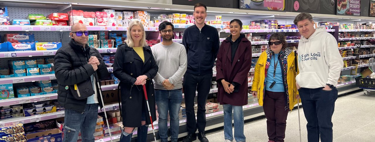 Sight Loss Council members stood in a line with a retailer facing the camera and smiling in a supermarket where they have been assessing the accessibility of product packaging. Three members are holding navigation canes.