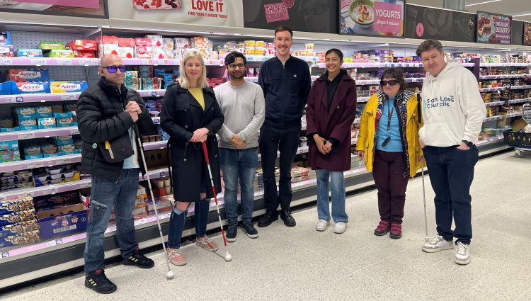 Sight Loss Council members stood in a line with a retailer facing the camera and smiling in a supermarket where they have been assessing the accessibility of product packaging. Three members are holding navigation canes.