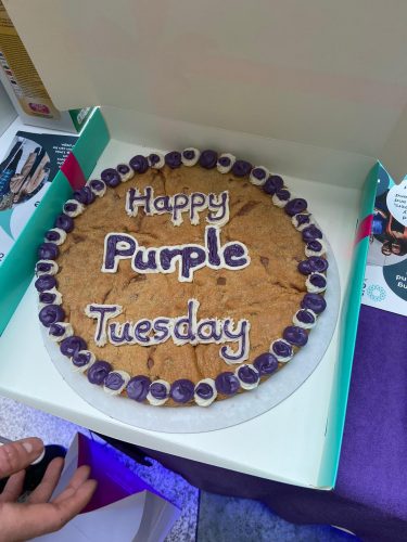 Image shows a large cookie with 'Happy Purple Tuesday' in purple and white icing in a cardboard box. 