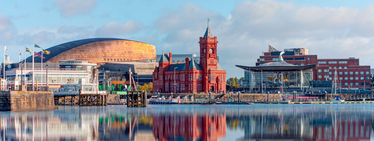 Panoramic view of Cardiff Bay in Cardiff, Wales. The view is of a lake with the red Pierhead building in the background, and buildings, reflecting in the water. Blue sky dotted with clouds.