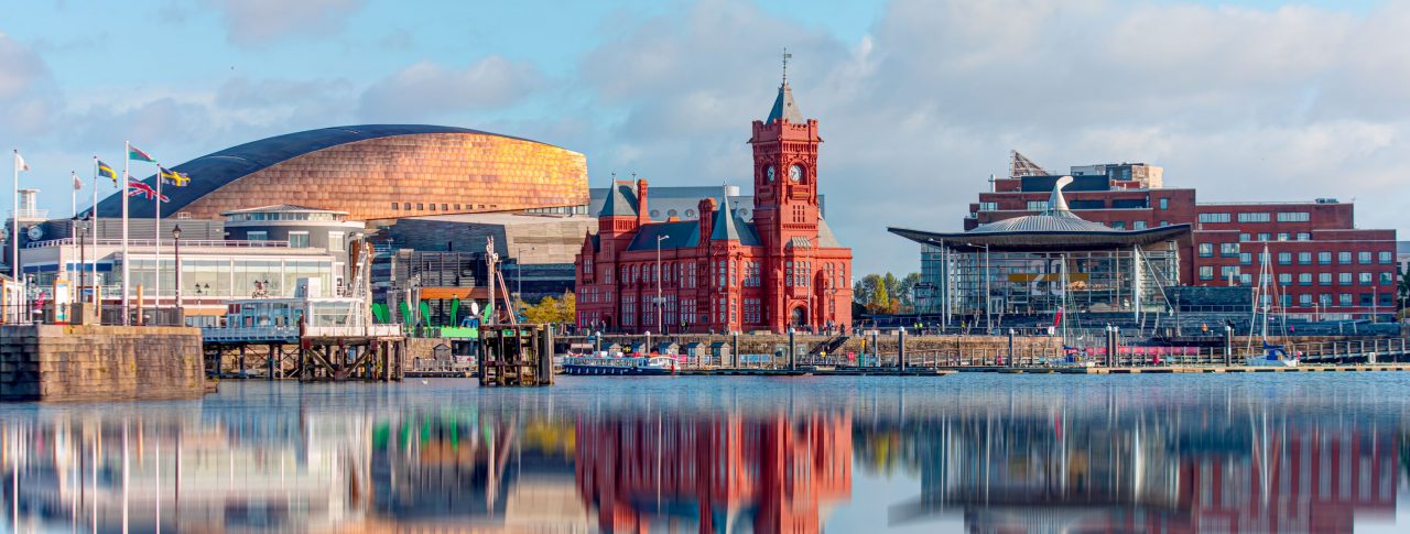 Panoramic view of Cardiff Bay in Cardiff, Wales. The view is of a lake with the red Pierhead building in the background, and buildings, reflecting in the water. Blue sky dotted with clouds.