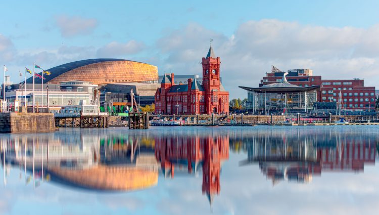 Panoramic view of Cardiff Bay in Cardiff, Wales. The view is of a lake with the red Pierhead building in the background, and buildings, reflecting in the water. Blue sky dotted with clouds.