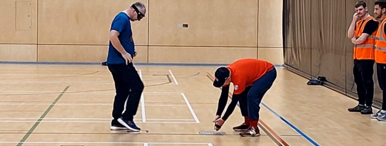 There are four people in a sports hall. A man in a blue top and black trousers has back facing a net and is standing in front of another man in a red top who is knelt a few feat away from him and placing a ball on the ground. There are two people in orange tabards standing against a black wall behind them.