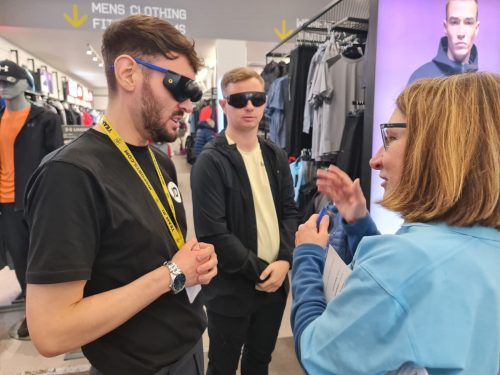 An SLC member speaking to two members of shop staff, both of whom are wearing sim specs. They are within a clothing shop. 