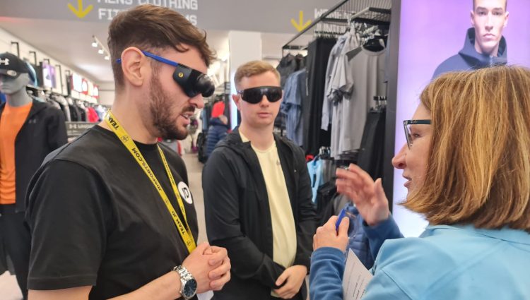 Female Sight Loss Council member talking to retails about accessible retail and the experiences of shoppers. The retailers are wearing sim specs (simulation glasses which simulate eye conditions) and are stood in a clothes store, wearing yellow lanyards as part of an event.